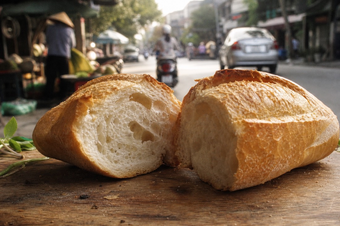 Split Hanoi banh mi baguette on a wooden board showing its dense crumb and hard crust in morning light
