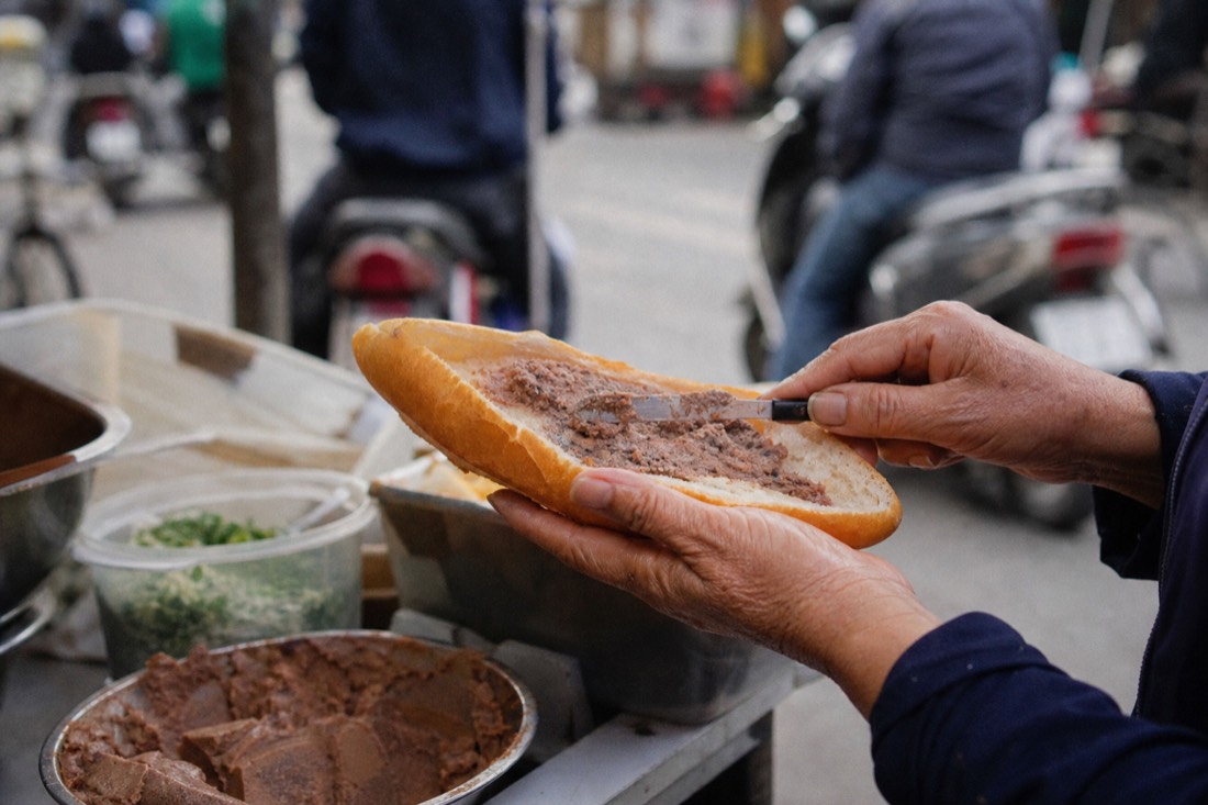 Vendor's hands spreading pate onto a freshly split baguette at a Hanoi street stall in close mid-shot