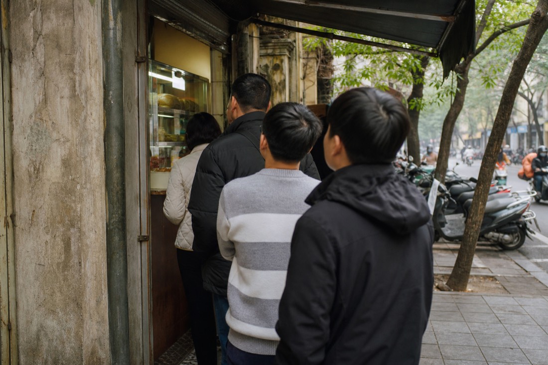 Short queue of customers waiting at a small banh mi stall window in Hanoi's Old Quarter in morning light