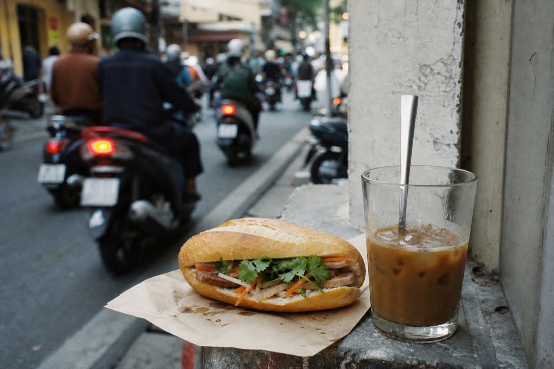 Banh mi sandwich and a glass of iced coffee resting on a narrow street ledge in Hanoi during the morning rush
