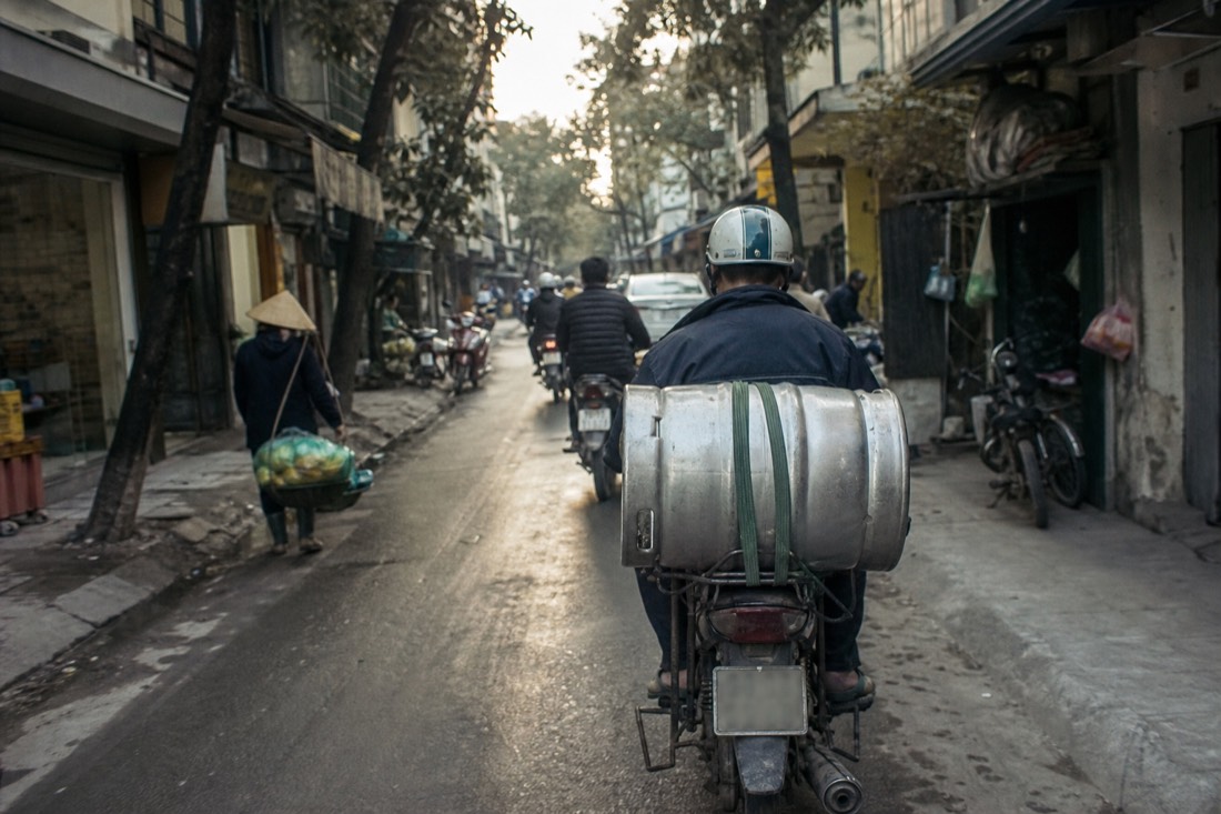 Steel beer keg strapped to the back of a motorbike navigating a narrow Hanoi street in early morning light