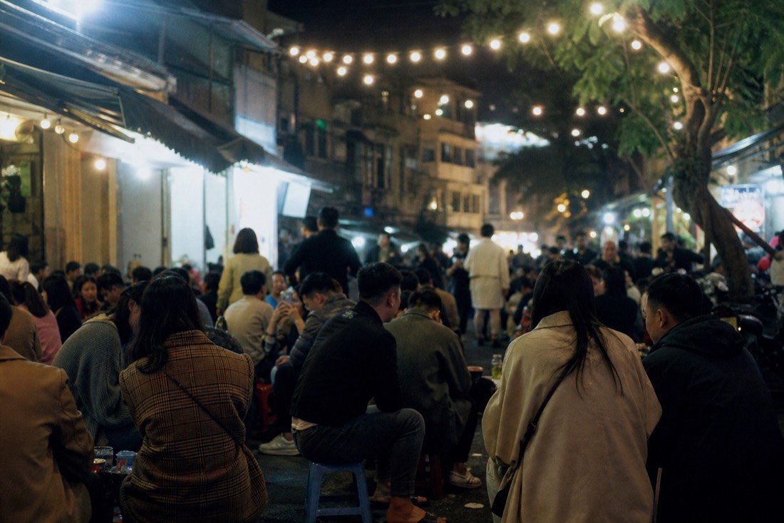 Busy street corner crowded with low plastic stools and small drink tables at night in Hanoi's Old Quarter under string lights