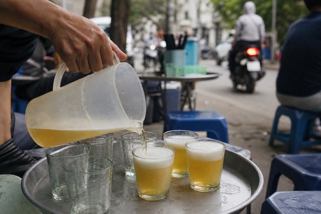 Vendor's hand pouring bia hoi from a jug into small glasses on a metal tray at a Hanoi street stall