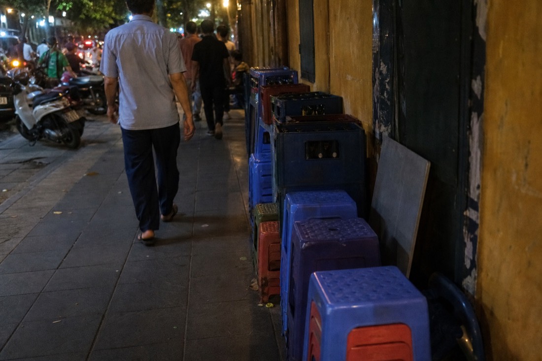 Stools and beer crates pulled tightly against a building wall on a narrow Hanoi pavement, leaving a clear walking path in evening light
