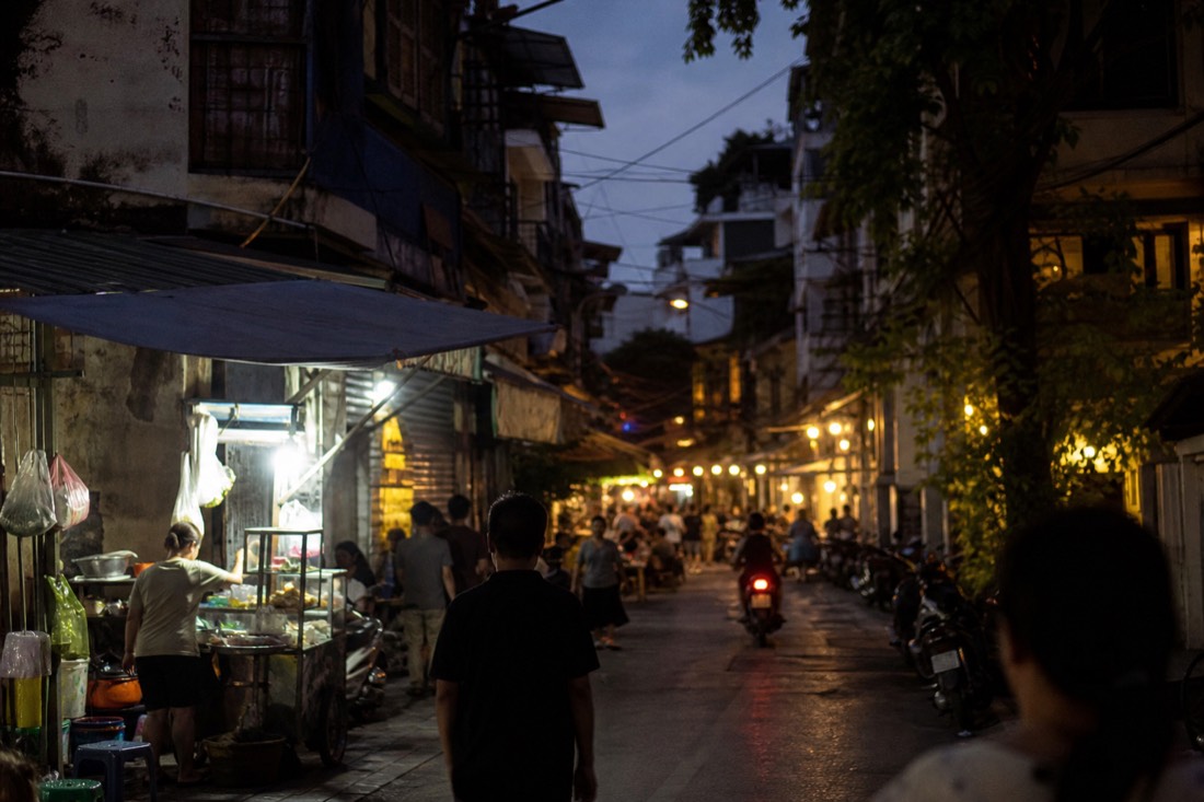 Narrow Old Quarter street at dusk with food stall lights on one side and the glow of a bia hoi corner further down in Hanoi