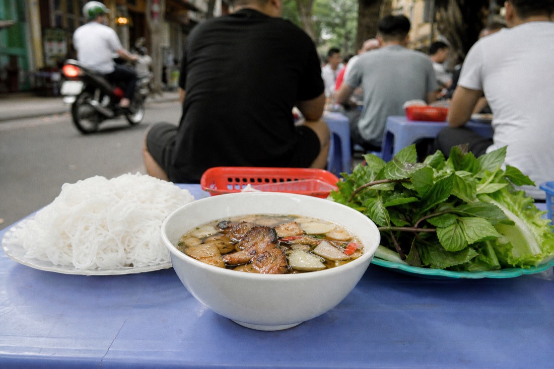 Bun cha dipping sauce bowl with grilled pork beside a plate of rice noodles and a basket of fresh herbs on a street food table in Hanoi