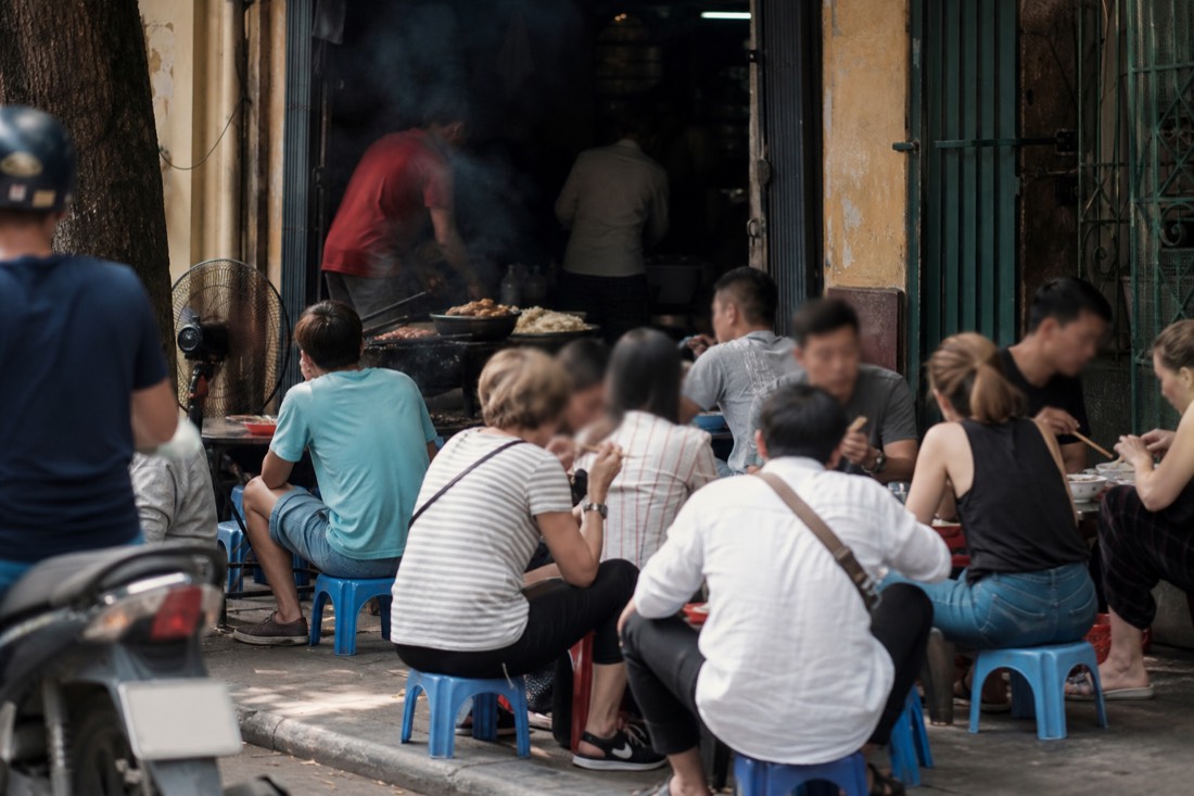 Busy bun cha shopfront in Hanoi's Old Quarter at midday with low plastic stools filled and smoke rising from a charcoal grill just inside the doorway