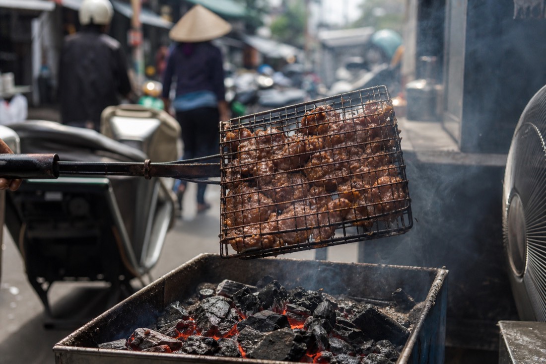 Hinged metal grill basket loaded with pork being fanned over glowing charcoal embers at a Hanoi street stall