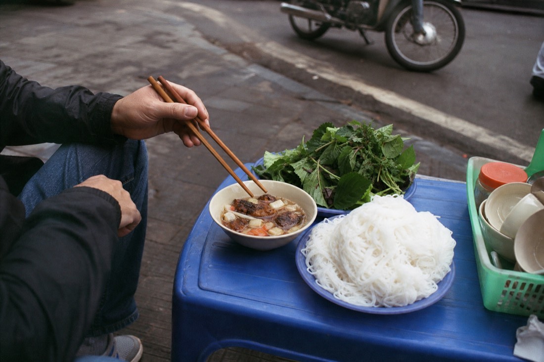 A lone diner's hands using chopsticks over a bowl of bun cha dipping sauce at a low outdoor table in Hanoi
