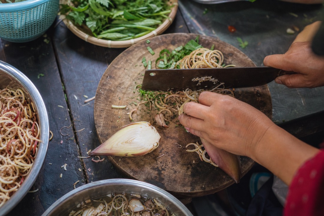 Thinly sliced banana flower and herbs being prepared on a wooden chopping board in a Hanoi kitchen, close overhead shot