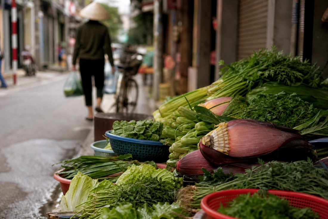 Vendor's market stall in Hanoi displaying fresh vegetables, herbs and a banana flower for sale in morning light