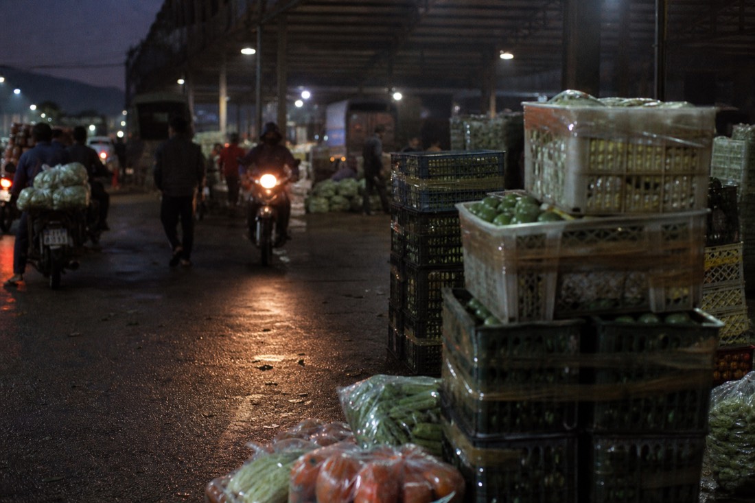 Headlamps and dim overhead lights illuminating stacked produce crates at a predawn wholesale market in Hanoi