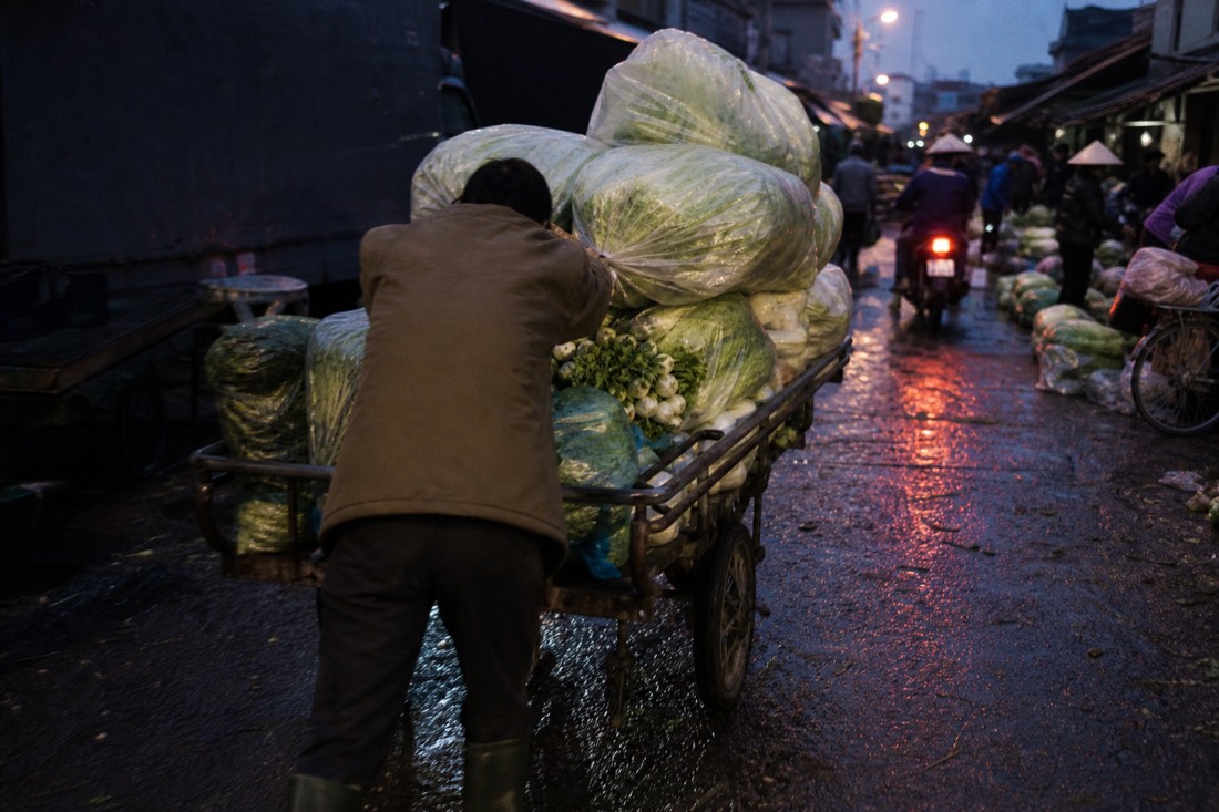Heavily loaded handcart of produce being pushed through a narrow lane at a predawn Hanoi wholesale market