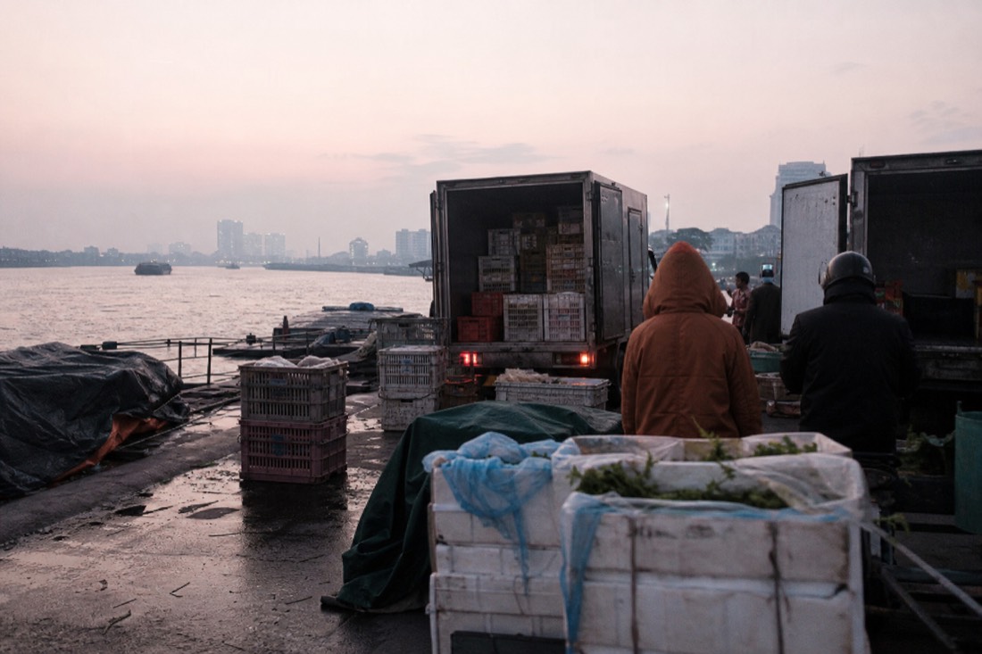 First grey light of dawn breaking over a riverside wholesale market in Hanoi with parked delivery trucks and stacked crates