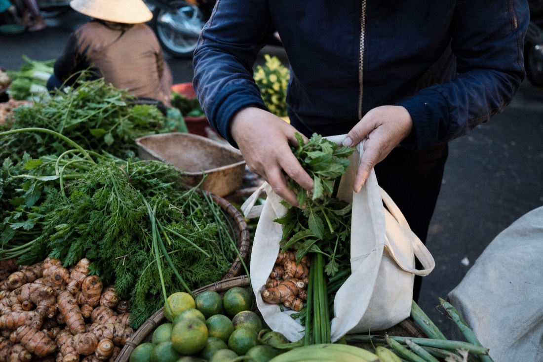 Fresh herbs, limes and turmeric roots being placed into a cloth shopping bag at a Hanoi market stall in early morning light