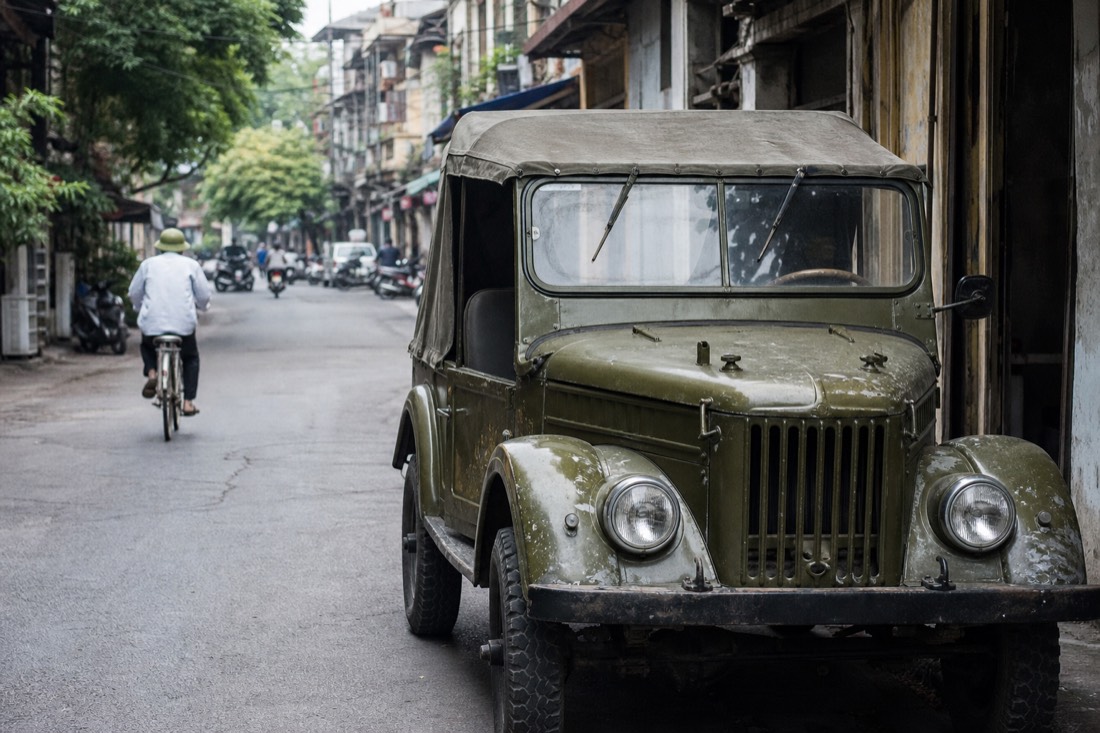 Vintage olive-green GAZ-69 army jeep parked empty on a narrow Hanoi Old Quarter street in daylight