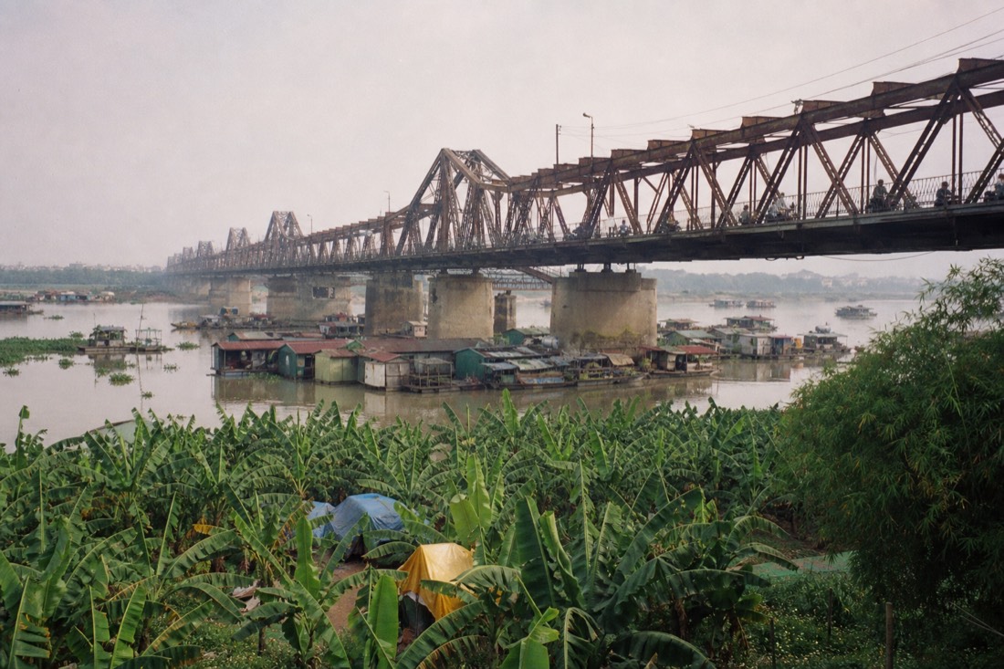 Old Long Bien rail bridge over the Red River in Hanoi with a floating village and banana groves visible below in hazy daylight