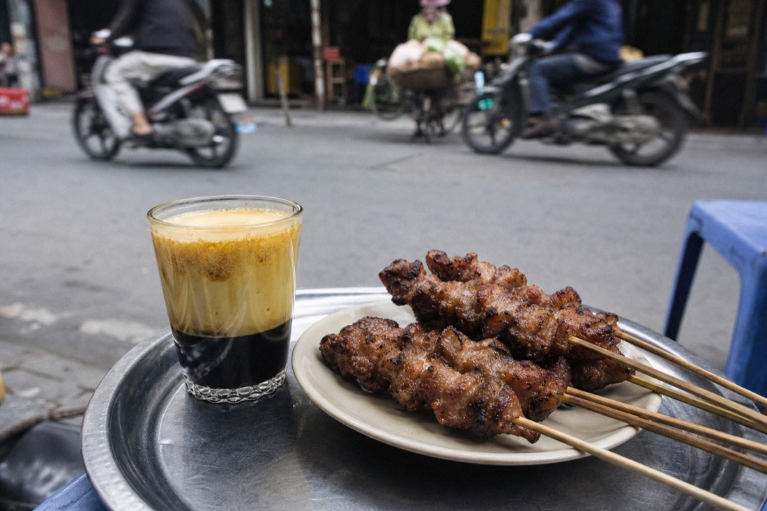 Grilled pork skewers and a glass of Vietnamese egg coffee arranged on a metal tray at a Hanoi street food stall in daylight