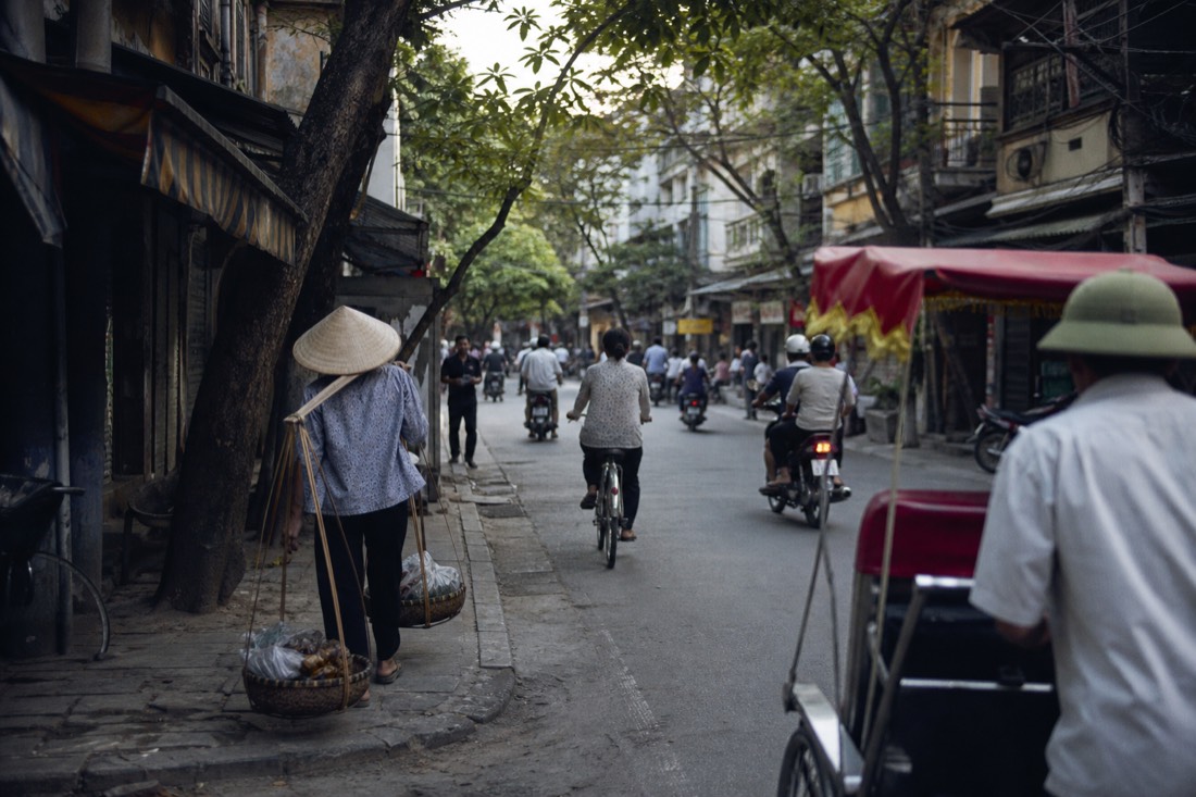 Hanoi Old Quarter street with pedestrians, bicycles and motorbikes moving past narrow shopfronts at midday