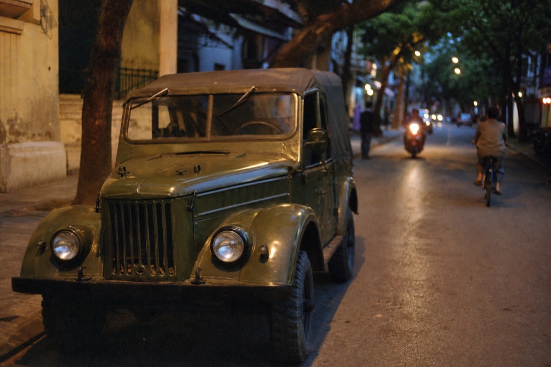 Vintage olive-green GAZ-69 jeep parked empty on a quiet Hanoi street at dusk with warm streetlight glow