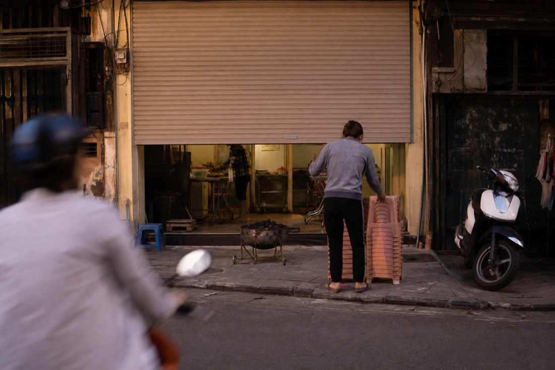 Shopfront shutter half-lowered at dusk with a charcoal brazier and stacked plastic stools being set up on the pavement in Hanoi's Old Quarter