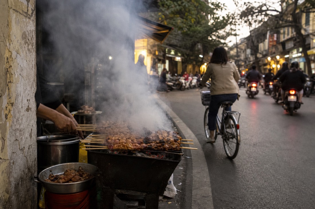 Street vendor's charcoal grill loaded with pork skewers on a narrow Hanoi Old Quarter corner at early evening with smoke rising