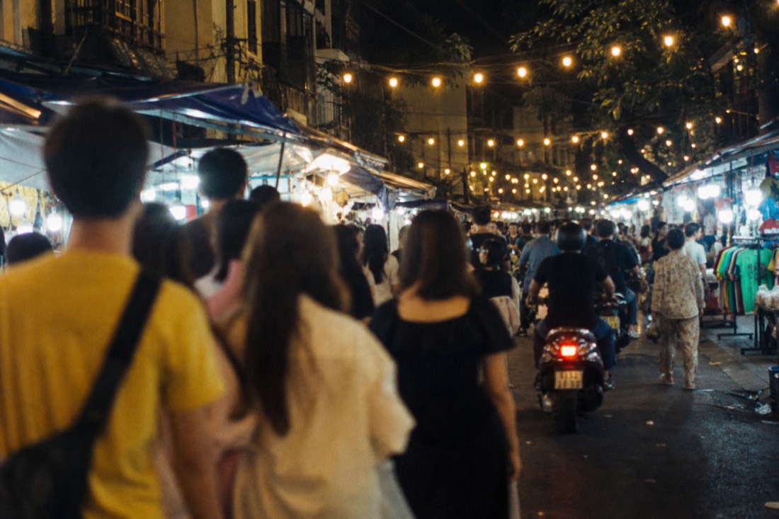 Crowded pedestrian night market street in Hanoi's Old Quarter with rows of small stalls under string lights on a weekend evening