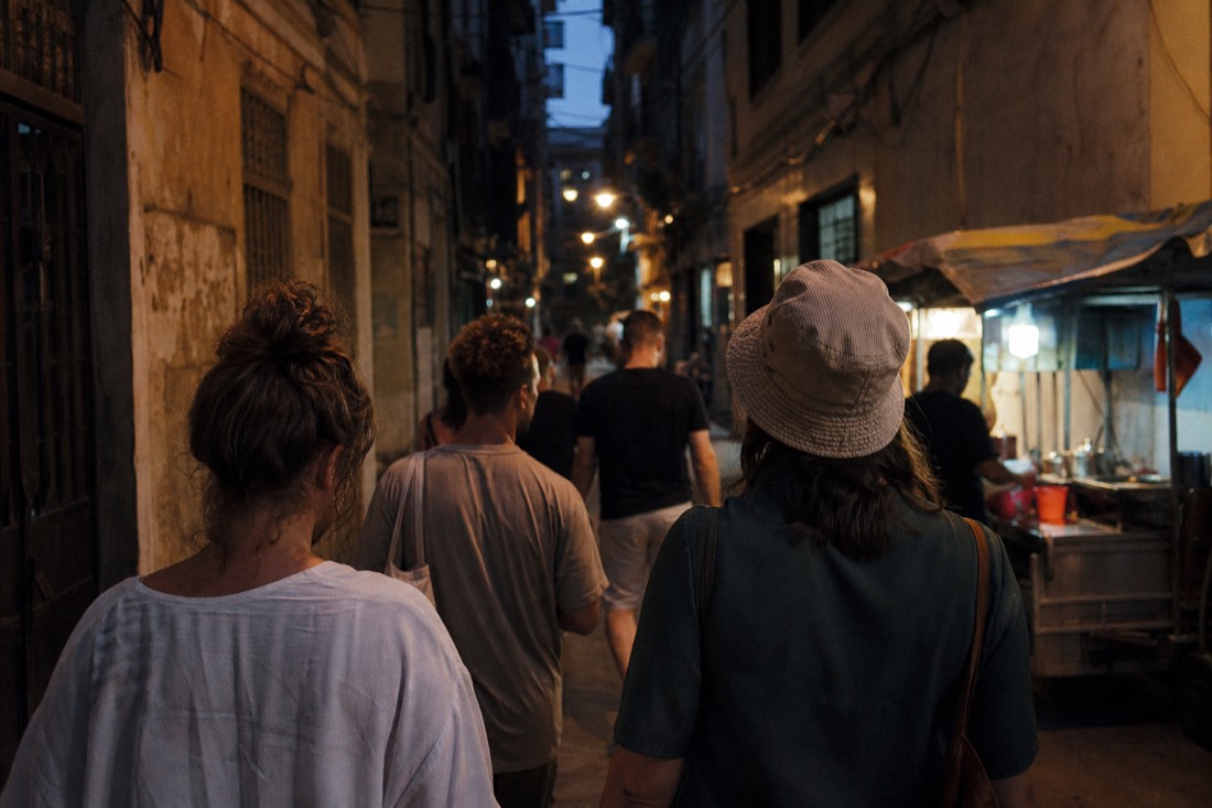 Small group of travellers walking together through a narrow lit Old Quarter alley at dusk in Hanoi, seen from behind