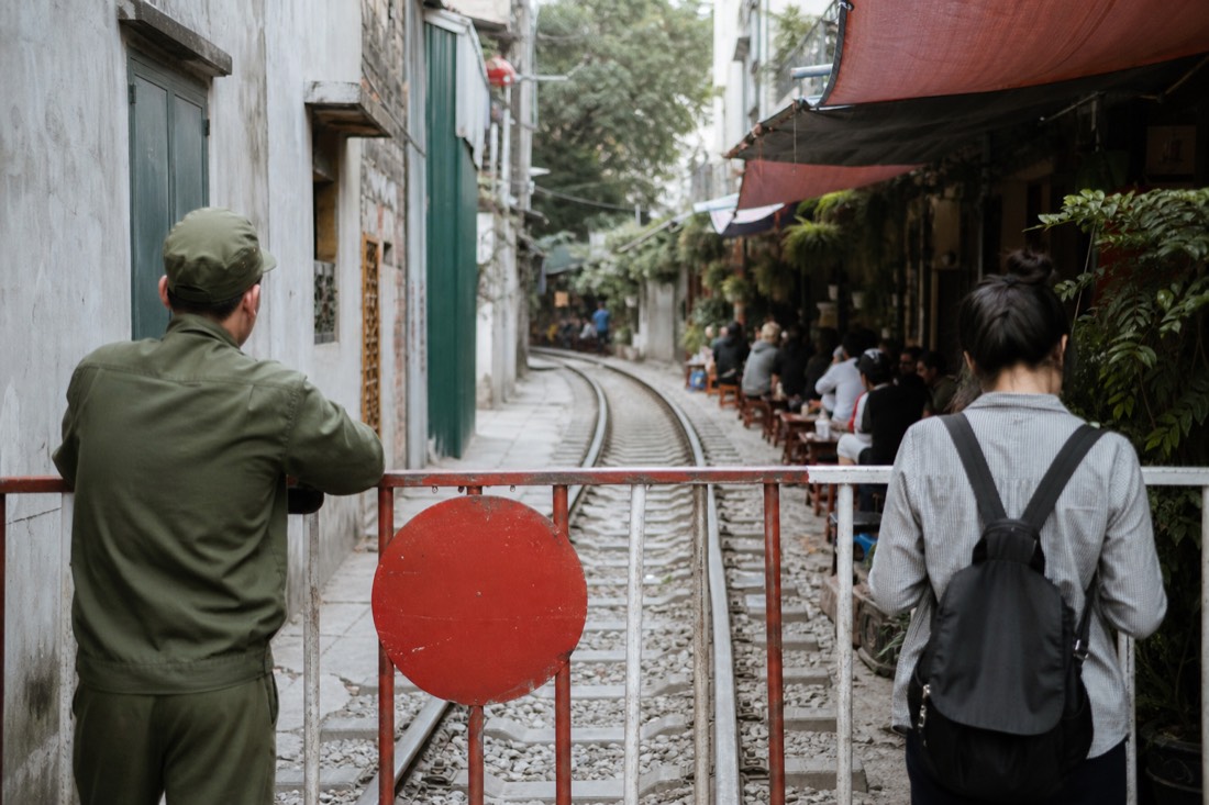 Barricaded Hanoi Train Street entrance with a metal gate and a track-side cafe with stools visible just beyond it in daytime