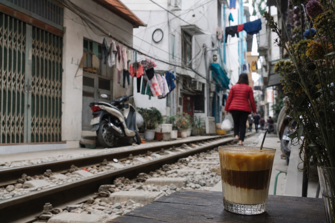 Glass of layered Vietnamese egg coffee on a small table beside railway tracks with laundry and cables overhead in Hanoi