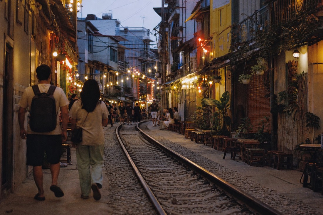 Train Street corridor at dusk with warm string lights glowing above the tracks and cafe stools along the walls in Hanoi