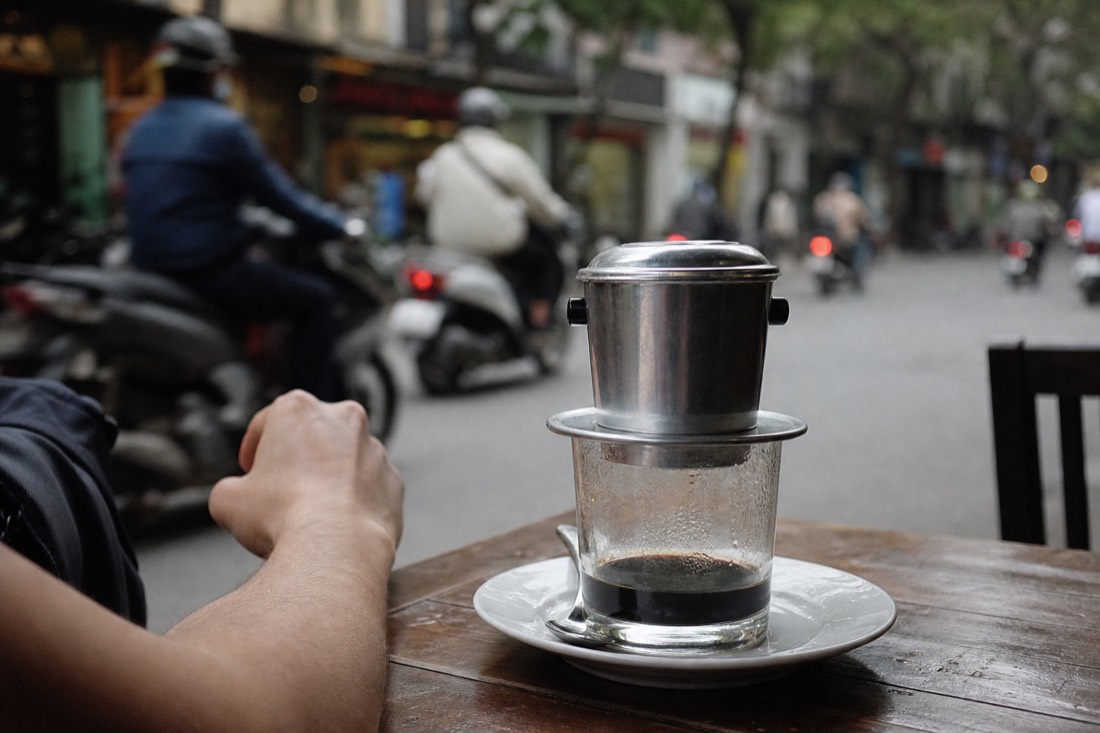 Metal phin filter sitting atop a glass on a saucer at a pavement cafe table in Hanoi with a blurred street behind