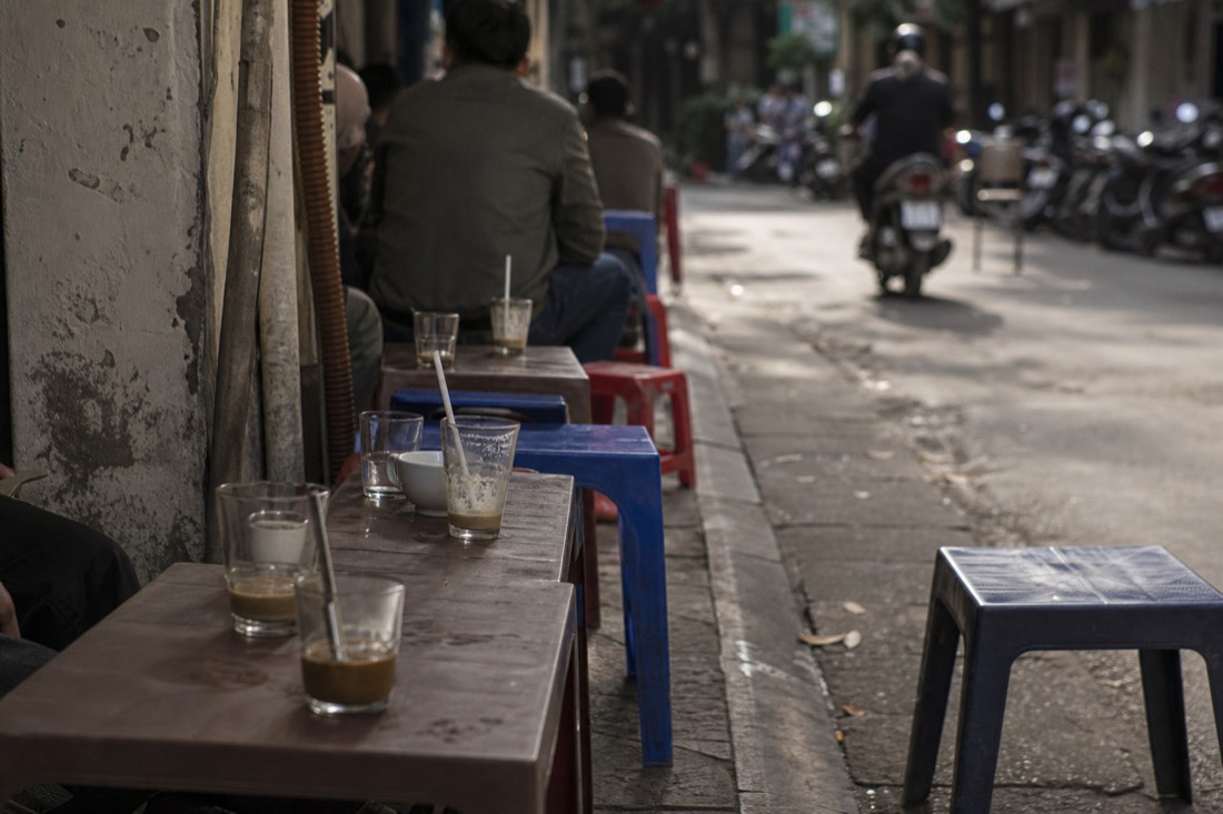 Row of small stools and low tables at a pavement coffee stall in Hanoi with cups left mid-sip in midday light