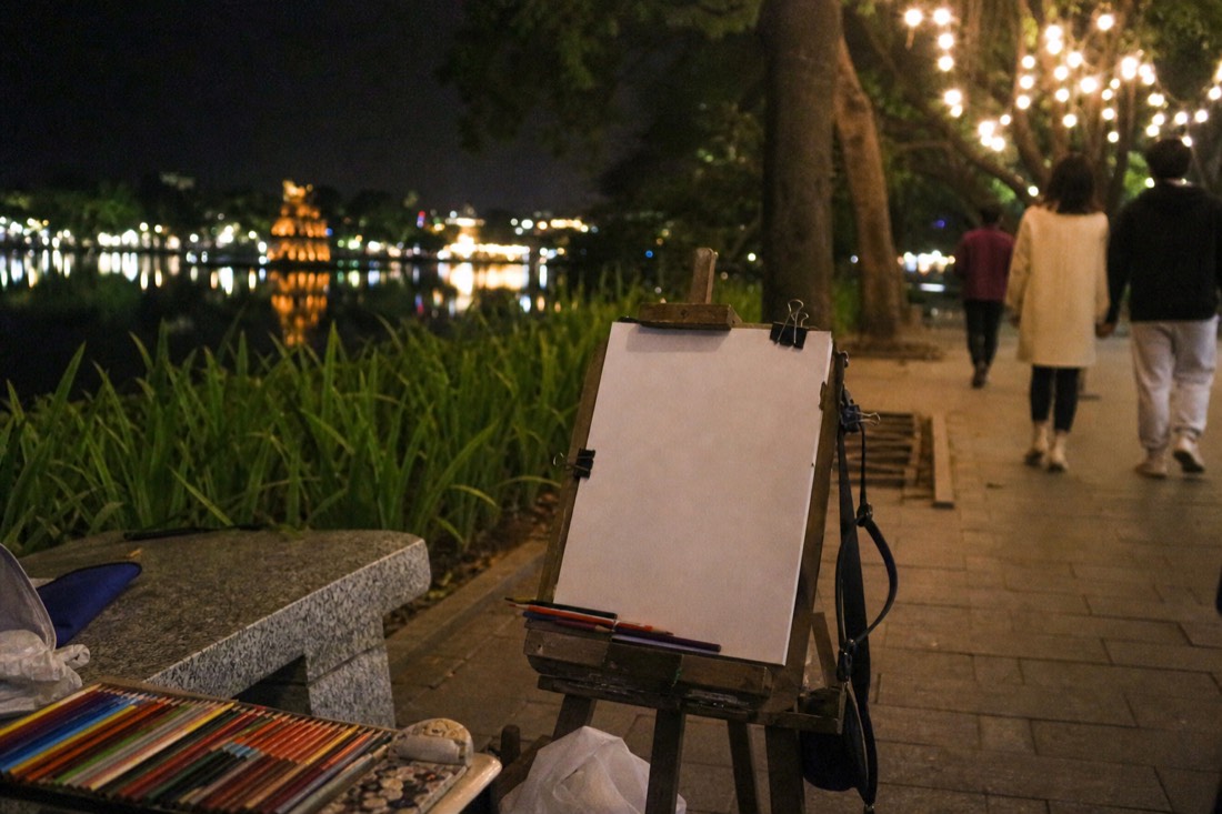 Portrait artist's easel and colored pencils set up along a lakeside promenade at night in Hanoi with string lights in the background