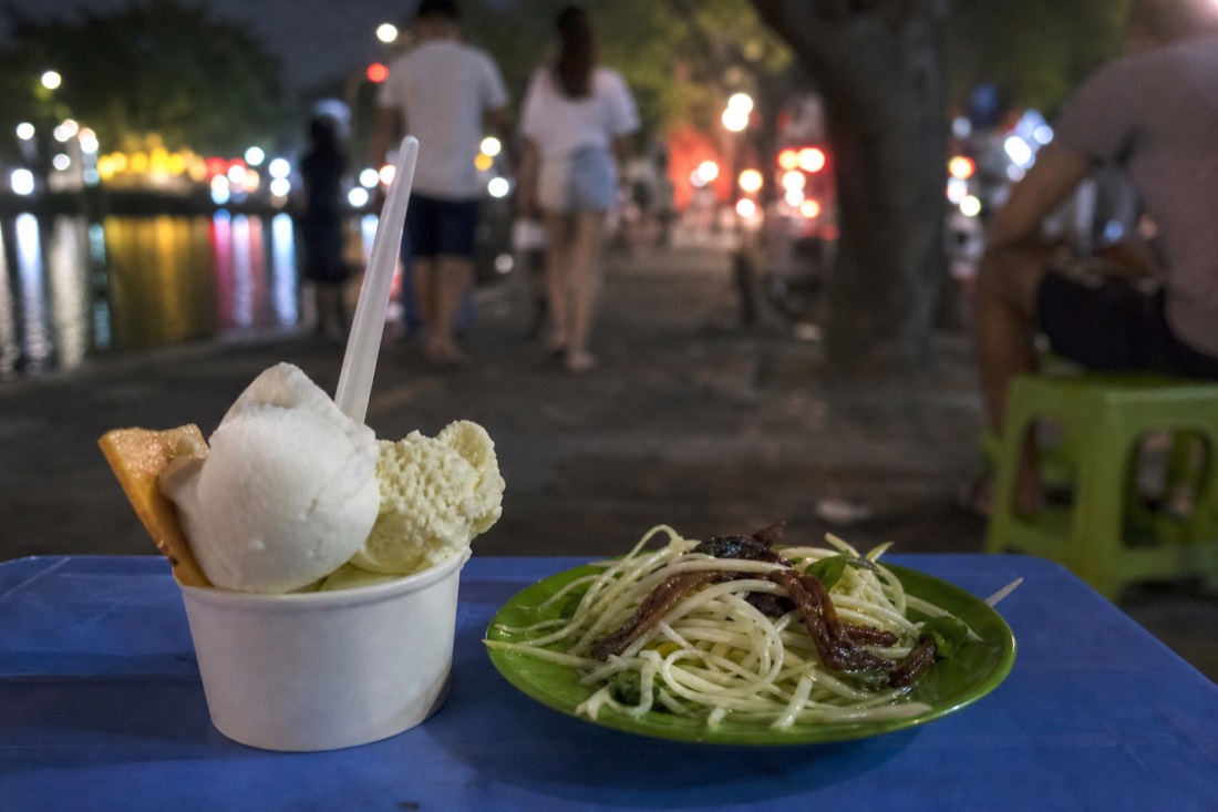 Cup of Trang Tien-style ice cream and a small plate of green papaya salad with beef jerky on a low table beside a lakeside promenade at night in Hanoi
