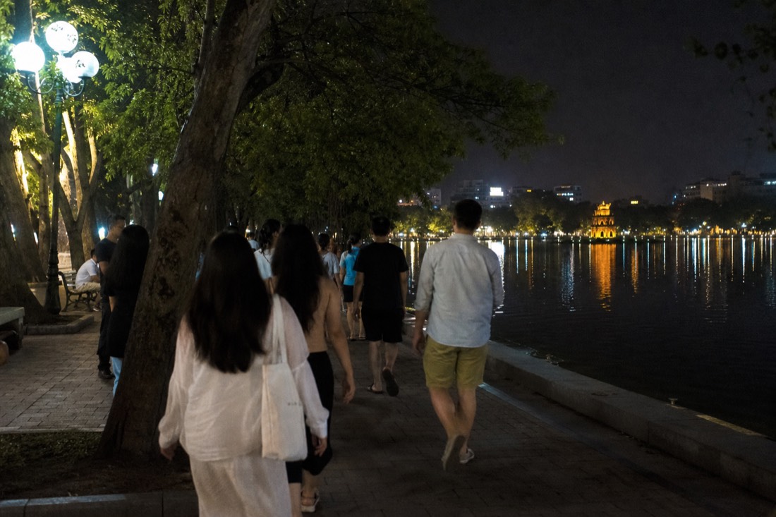 Hoan Kiem Lake promenade at night with warm lights reflecting on the water and strolling crowds in the distance