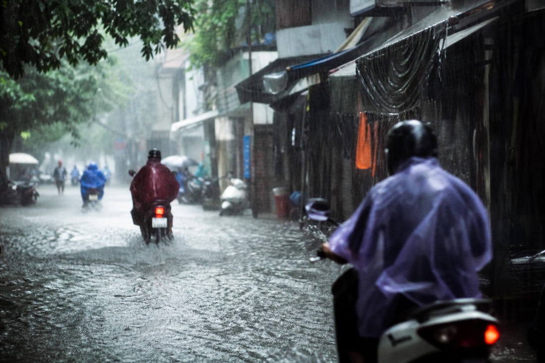 Sudden late-afternoon downpour flooding a narrow Hanoi street with motorbikes splashing through