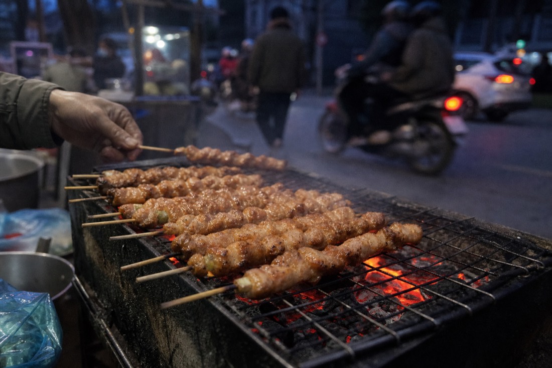 Charcoal grill glowing at dusk with pork skewers cooking at a Hanoi street stall in cool evening air