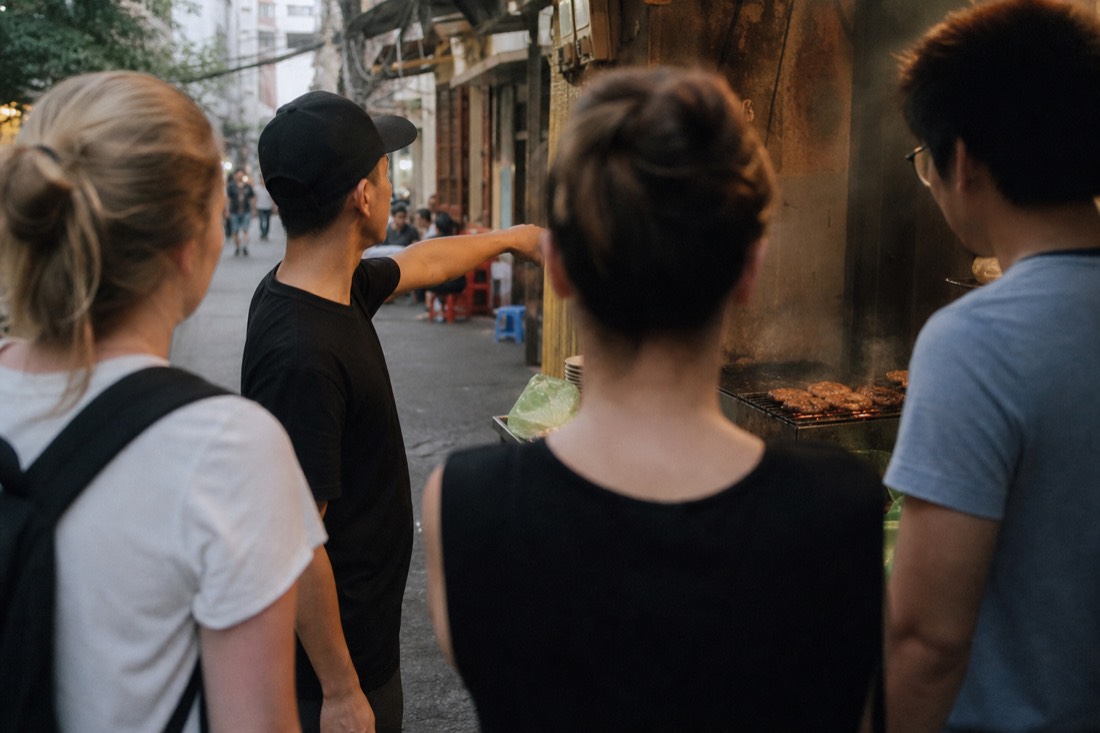 Guide gesturing toward a bun cha grill stall while a small group watches in a Hanoi Old Quarter alley at early evening
