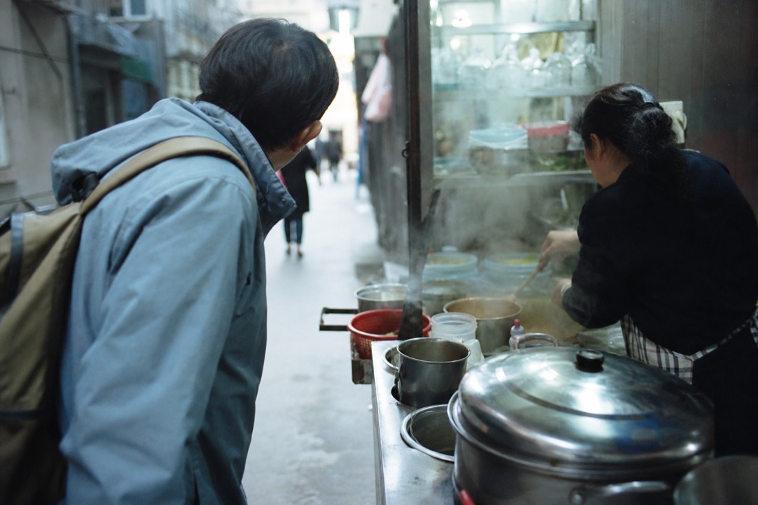 Local guide speaking with a street food vendor over a counter of simmering pots in a Hanoi alley kitchen