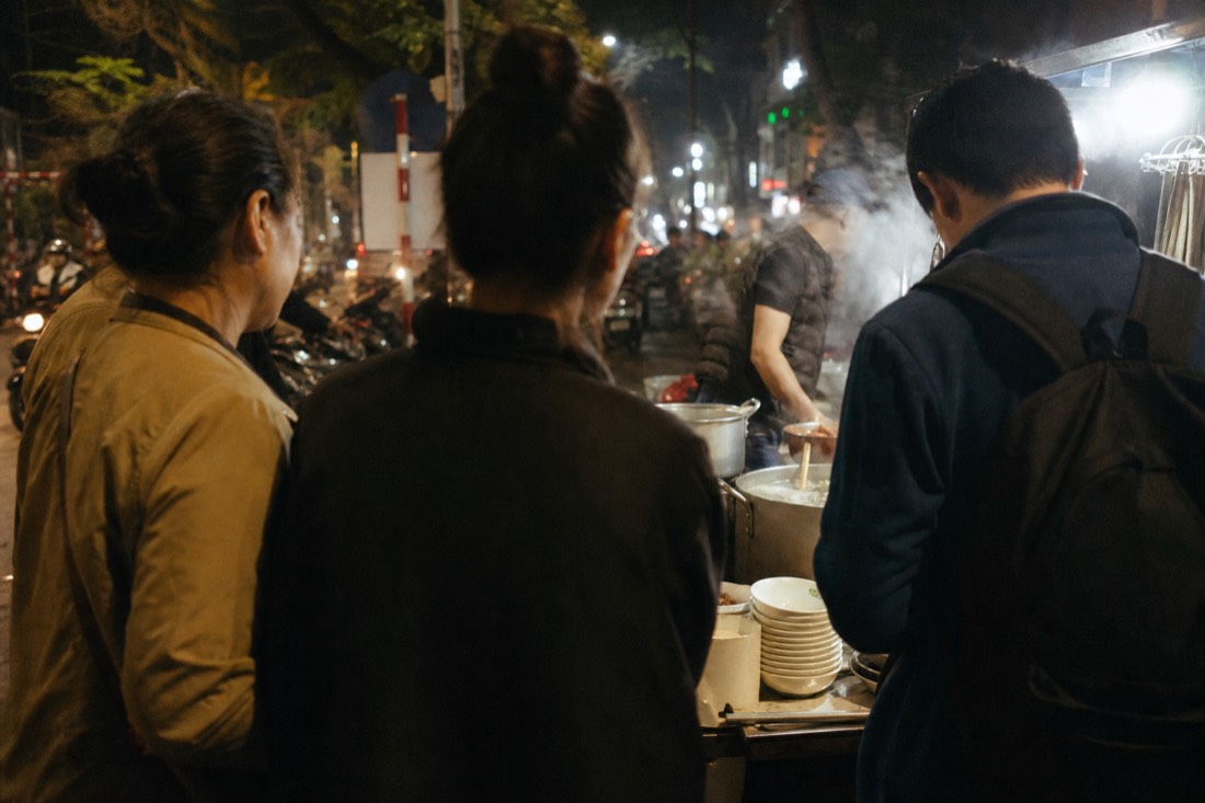 Guide and small group standing at a busy Hanoi street food stall pointing at simmering pots