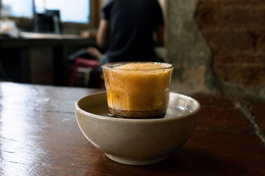 Cup of Hanoi egg coffee sitting in a bowl of hot water on a worn wooden cafe table