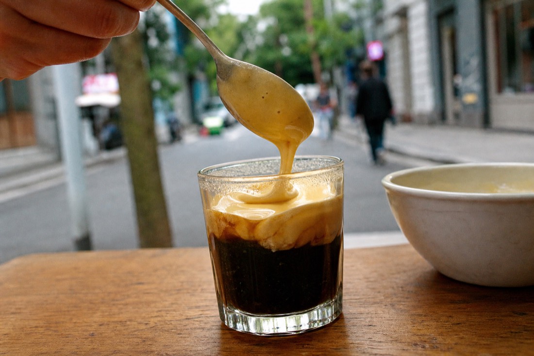 Close-up of whipped egg yolk cream being spooned over dark robusta coffee in a small glass