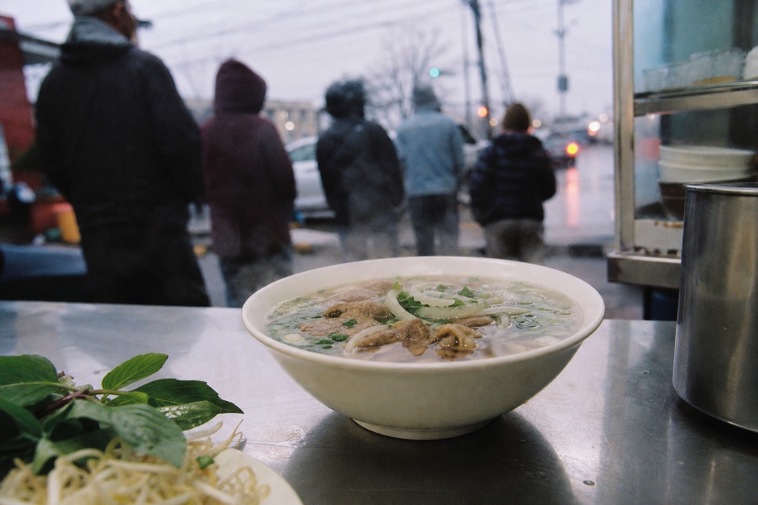 Steaming bowl of pho on a counter at a Hanoi street stall at dawn with a queue of customers blurred behind