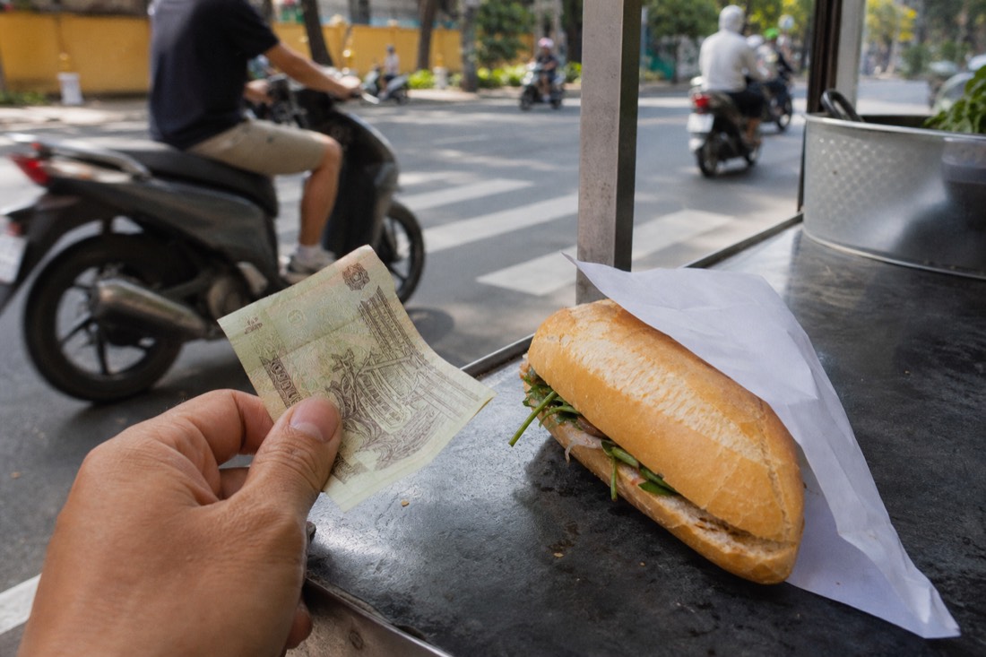 Hand holding a small Vietnamese banknote beside a plain banh mi wrapped in paper on a street cart