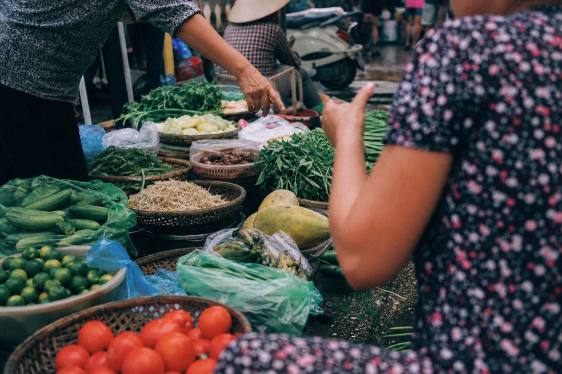 Wet market stall piled with fresh produce and a vendor's hand pointing toward a basket