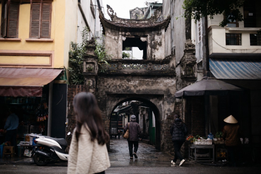 Old stone temple gate wedged between narrow shophouses in Hanoi's Old Quarter market street