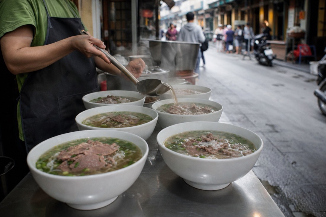 Steaming bowls of beef pho lined up on a stainless counter at a Hanoi Old Quarter pho stall in early morning
