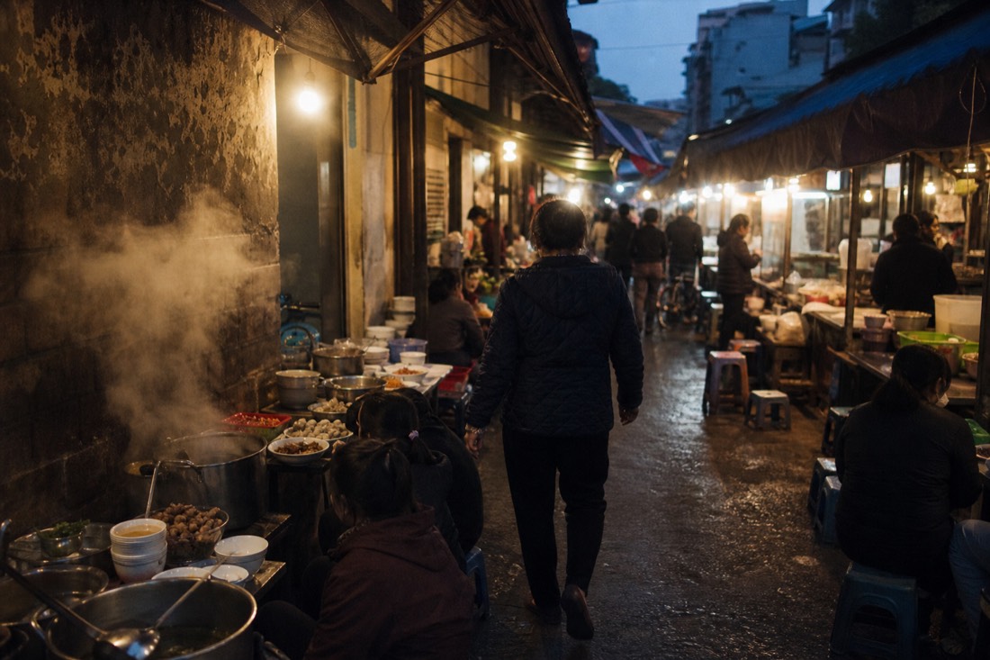 Crowded evening food alley beside Dong Xuan Market in Hanoi with counters and low stools