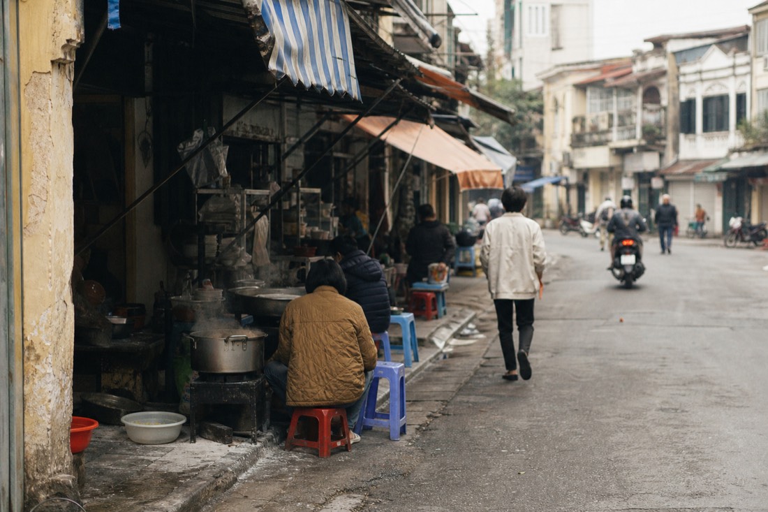 Cluster of open-front food stalls with steaming pots along a narrow Hanoi Old Quarter street corner at midday